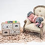 baby, newborn, sleeping, armchair, vintage_furniture, checkered_clothing, cap, pants, decorative_blocks, name_blocks, drawer_chest, rug, beige, white_background, cozy, portrait, infant, peaceful, studio_photo, soft_lighting