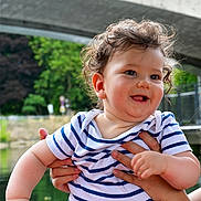 Nelson participe au concours pour gagner de l'argent avec cette photo : baby, child, curly_hair, striped_shirt, smiling, outdoor, pond, bridge, greenery, hands, person, happy, cute, portrait, nature, water, summer, holding, infant, playful