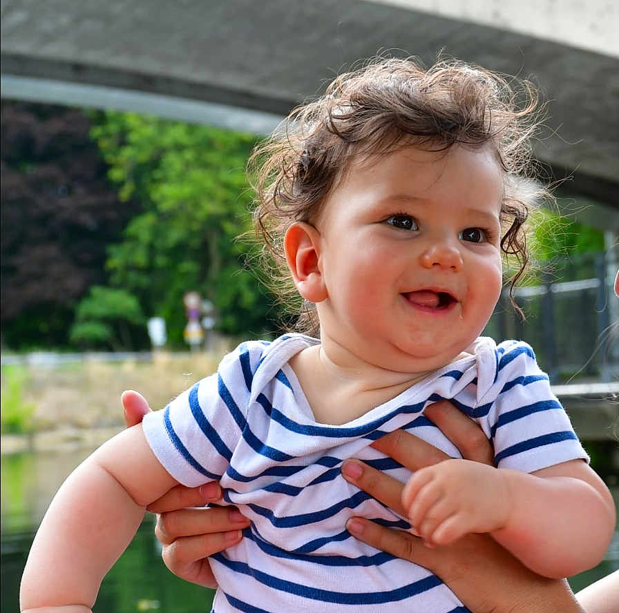 Nelson participe au concours pour gagner de l'argent avec cette photo : baby, child, curly_hair, striped_shirt, smiling, outdoor, pond, bridge, greenery, hands, person, happy, cute, portrait, nature, water, summer, holding, infant, playful