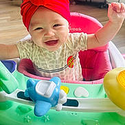 Laylah is registered to the contest to win money with this photo: baby, infant, smiling, red_turban, highchair, toy_plane, play_center, joyful, teeth, chubby_cheeks, onesie, leopard_print, pink_seat, wooden_floor, indoor, hand_raised, close_up, portrait, feeding_seat, cute