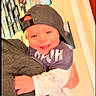 baby, child, infant, smiling, portrait, indoor, baseball_cap, backward_cap, onesie, clothing, hugging, adult, family, wall_art, photo_frames, home, warm_lighting, happy, sitting, close_up