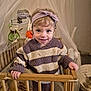 baby, baby_toys, blanket, child, clothing, crib, cute, happy, headband, indoor, mobile, net_canopy, nursery, person, portrait, smiling, striped_clothing, sweater, toddler, wooden_furniture