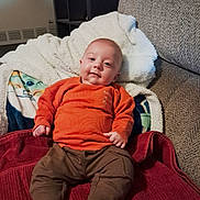 Noah joined the competition — help win amazing prizes! baby, blanket, brown_pants, child, couch, cozy, cute, fabric, footwear, home, indoor, infant, orange_sweater, person, portrait, relaxed, seated, smile, soft_toys, warm_clothing