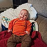 baby, blanket, brown_pants, child, couch, cozy, cute, fabric, footwear, home, indoor, infant, orange_sweater, person, portrait, relaxed, seated, smile, soft_toys, warm_clothing