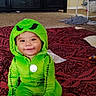 toddler, child, costume, green, smile, face, baby, sitting, blanket, pattern, indoor, carpet, toy, clothing, hood, pendant, furniture, home, baby_swing, cute