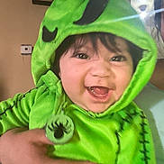 Promiseaity Flores joined the competition — help win amazing prizes! baby, child, costume, green_clothing, hood, smile, happy, face, hand, indoor, person, black_hair, light, lens_flare, wall, clock, cute, infant, holding, portrait