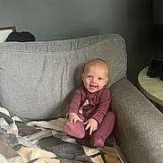 Bayleigh is registered to the contest to win money with this photo: baby, child, smiling, couch, blanket, indoors, onesie, happy, face, sitting, furniture, cozy, home, infant, person, cute, hands, foot, living_room, casual