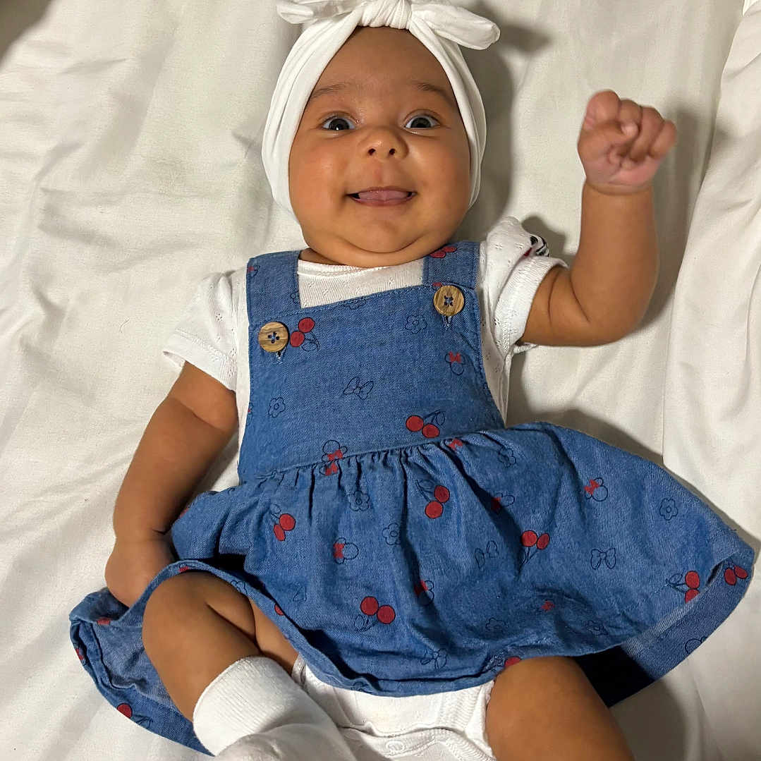 Amelia joined the competition — help win amazing prizes! baby, bed, blue_dress, bow, cherry_pattern, child, clothing, cute, happy, headband, indoors, infant, lying_down, person, playful, portrait, raised_arm, smiling, white_sheet, white_socks