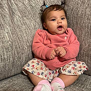 Isabella is registered to the contest to win money with this photo: baby, child, pigtails, pink_sweater, floral_skirt, socks, couch, indoor, cute, infant, smiling, hands, face, person, seated, comfortable, home, adorable, young, portrait