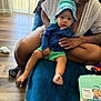 baby, child, woman, indoor, bucket_hat, blue_jacket, green_shorts, couch, wood_floor, vertical_blinds, hand, foot, person, seated, clothing, home, box, tattoo, skin, hat