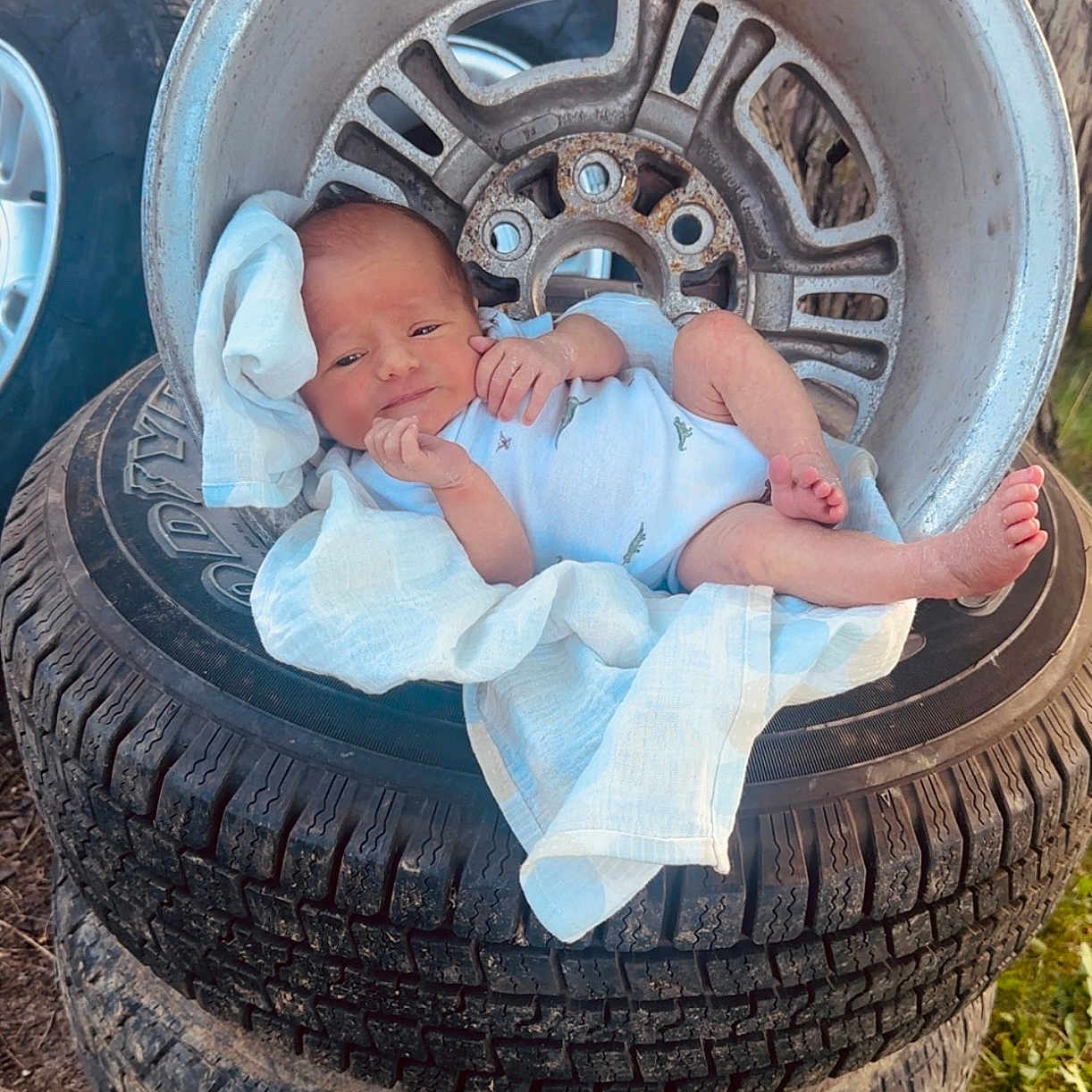 Jonah is registered to the contest to win money with this photo: baby, newborn, tire, wheel_rim, cloth, outdoor, grass, tree, car, vehicle, stacked_tires, baby_clothes, infant, person, foot, hand, face, posing, daylight, nature
