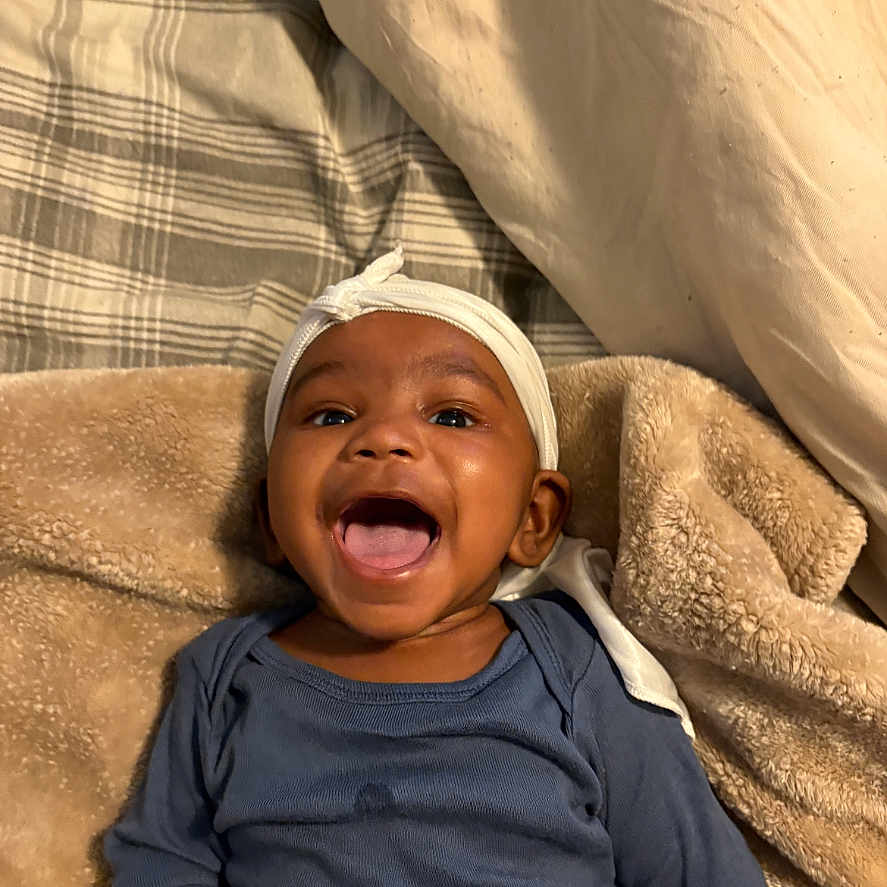 Idris is registered to the contest to win money with this photo: baby, blanket, child, clothing, cozy, cute, face, happy, headwrap, human, indoor, infant, lying_down, mouth_open, pillow, plaid, portrait, smiling, soft_texture, young_child