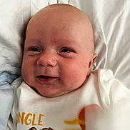 Lewis is registered to the contest to win money with this photo: baby, infant, face, smile, onesie, clothing, bed, blanket, cute, child, skin, head, portrait, sleepwear, young, lying_down, happy, indoors, soft, newborn