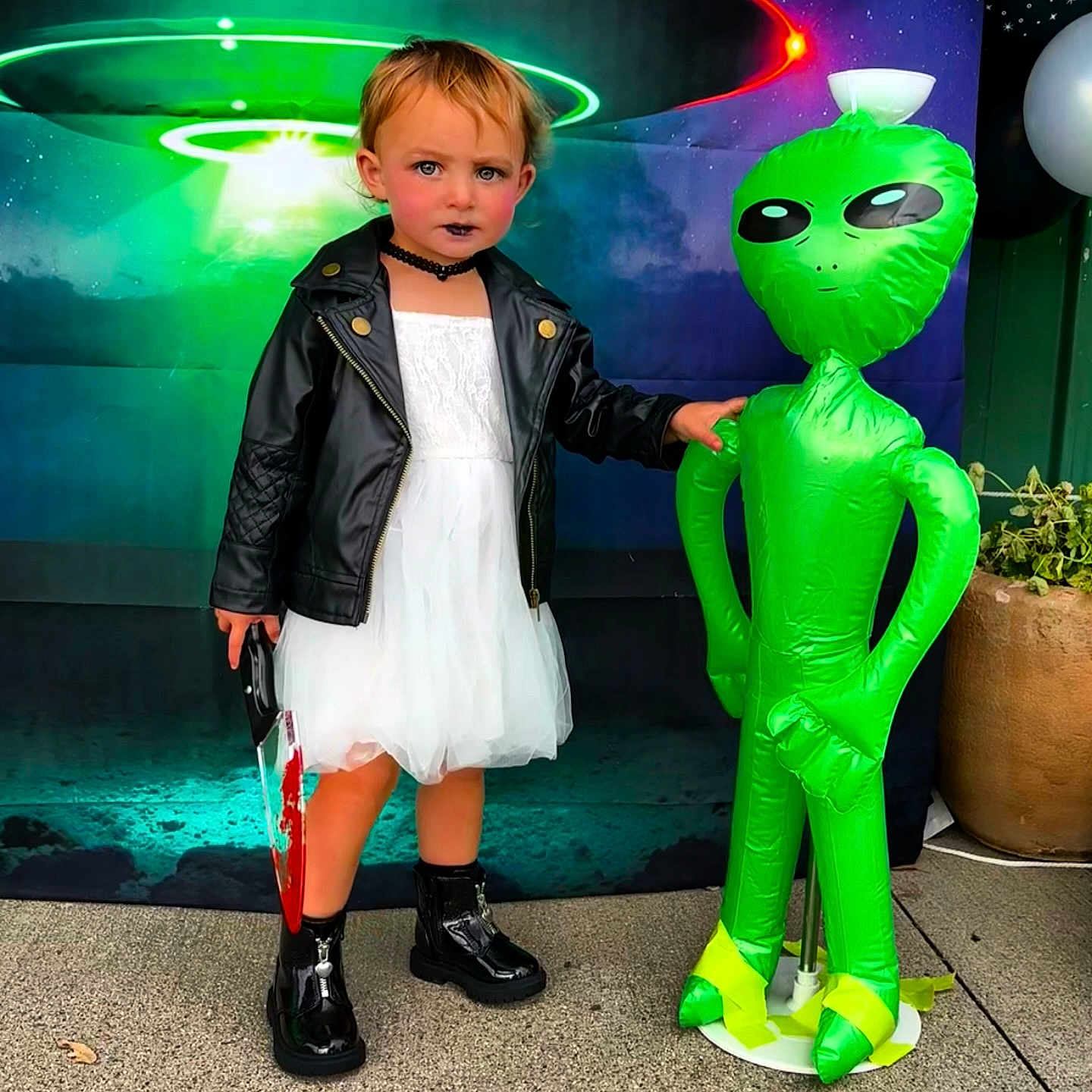 Devyn joined the competition — help win amazing prizes! child, kid, dress, leather_jacket, black_boots, inflatable_alien, toy_knife, halloween, costume, face_paint, makeup, background, ufo, green, white_dress, black_choker, outdoor, sidewalk, plant_pot, playful