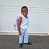 backpack, beads, black_shoes, blue_outfit, braids, casual_clothing, child, confident, daylight, garage_door, girl, glasses, outdoor, pavement, person, portrait, ready_for_school, smiling, standing, young