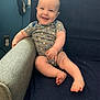 baby, smiling, onesie, couch, blue, armrest, home, infant, happy, face, legs, feet, wall, socket, plush, child, person, indoors, relaxed, seated