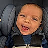 baby, blue_clothing, brown_hair, car_seat, child, close_up, face, gray_fabric, happy, harness, indoors, infant, joy, open_mouth, photography, portrait, safety_harness, seated, smiling, vehicle