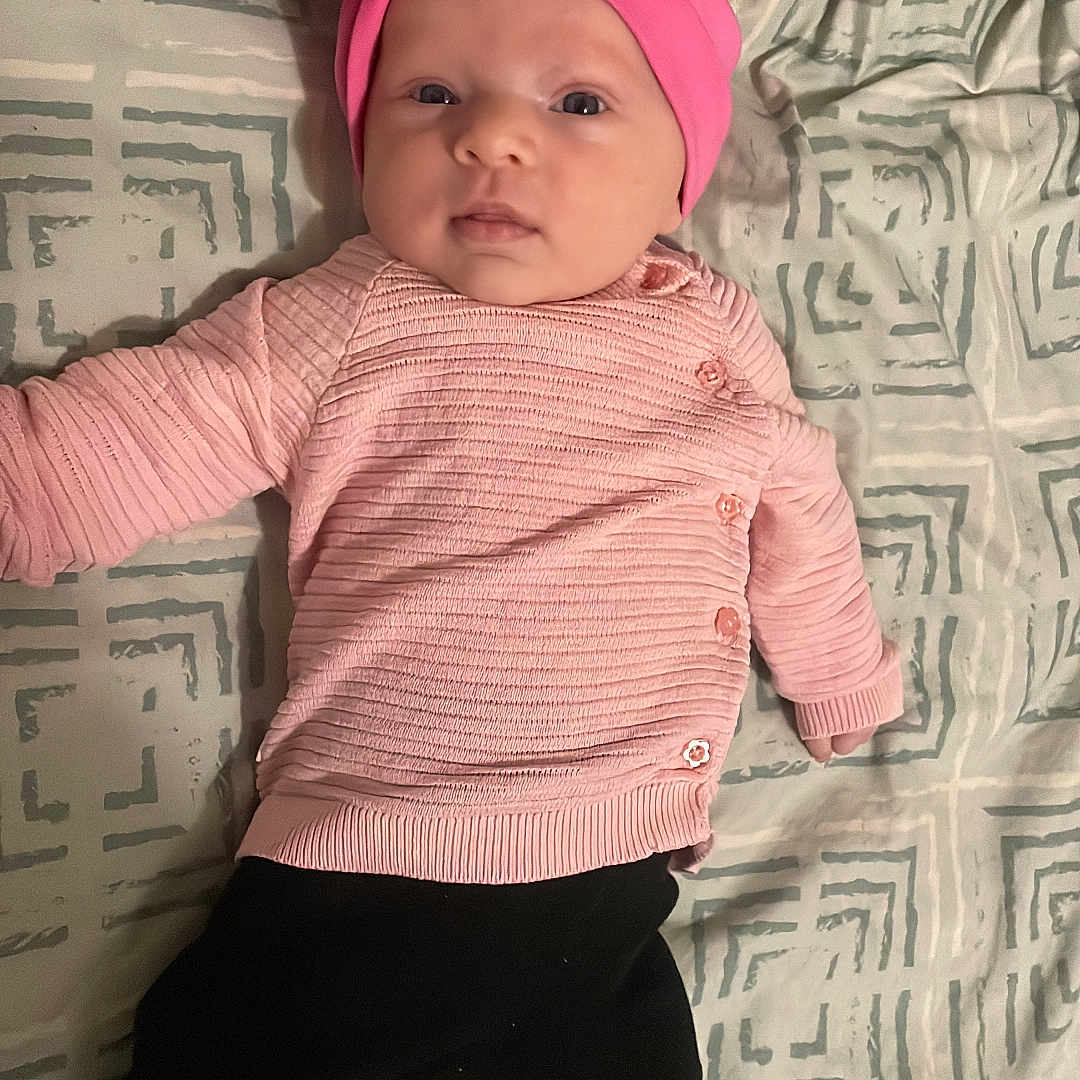 Jennah a rejoint le concours — aidez-le/la à gagner de superbes lots ! baby, baby_photo, bed, black_pants, blanket, cute, eyes, face, indoors, infant, infant_clothing, lying_down, newborn, one_person, patterned_bedding, pillow, pink_hat, pink_sweater, portrait, white_socks