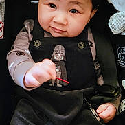 Ivy is registered to the contest to win money with this photo: toddler, child, baby, car_seat, black_clothing, darth_vader, cute, portrait, indoors, person, hair_ties, smile, holding_object, young_child, infant, seatbelt, fabric, face, looking_at_camera, casual
