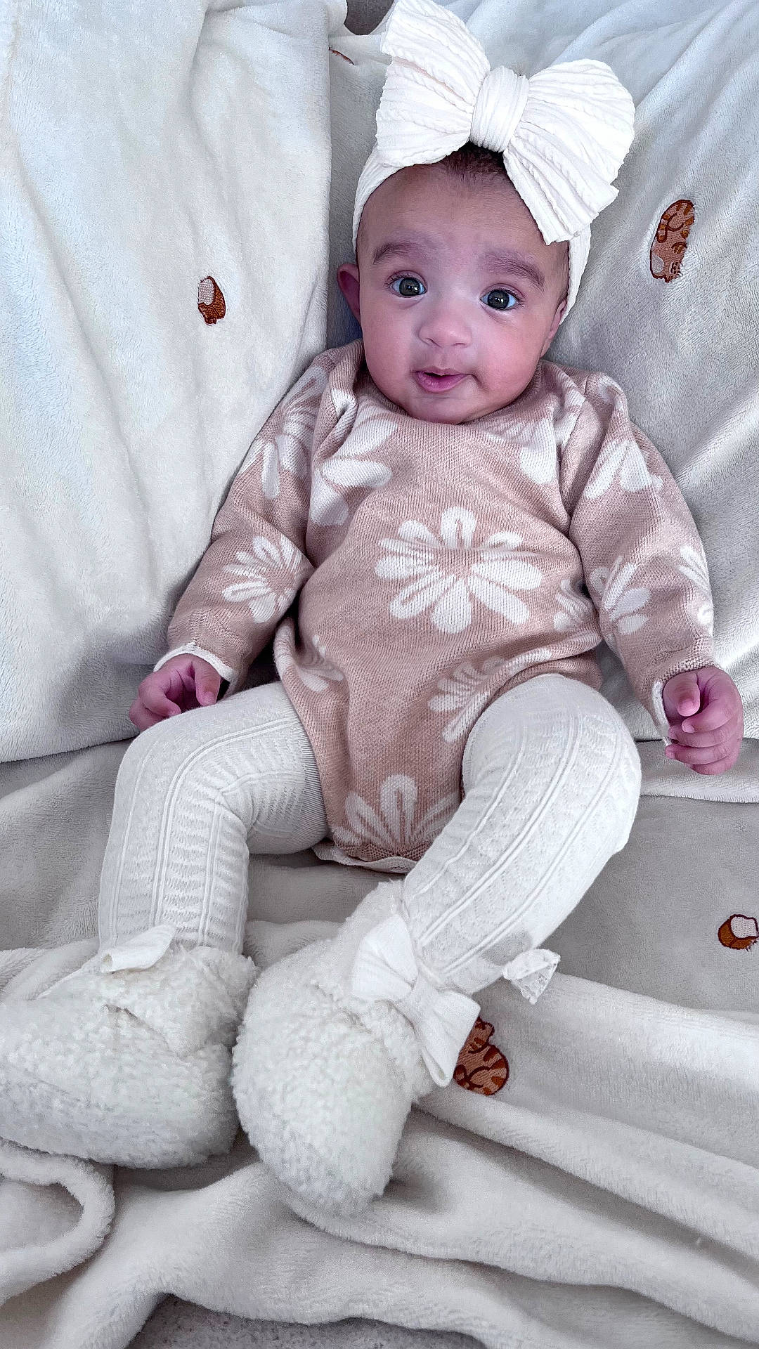 Heer is registered to the contest to win money with this photo: baby, blanket, bow, child, cozy, cute, face, floral_pattern, fuzzy_slippers, headband, indoor, infant, leggings, portrait, seated, slippers, smiling, soft_texture, sweater, white_leggings