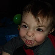 Andrew is registered to the contest to win money with this photo: baby, brown_hair, child, close_up, dim_lighting, eyes, face, gray_shirt, green_balloon, grinning, happy, indoor, infant, person, photograph, playful, portrait, red_clothing, sitting, smiling