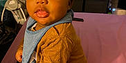 Messiah is registered to the contest to win money with this photo: baby, infant, child, sitting, indoor, clothing, bib, mustard_shirt, black_pants, pink_surface, television, cartoon, furniture, curious, cute, portrait, face, eyes, hair, room