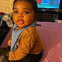 baby, infant, child, sitting, indoor, clothing, bib, mustard_shirt, black_pants, pink_surface, television, cartoon, furniture, curious, cute, portrait, face, eyes, hair, room