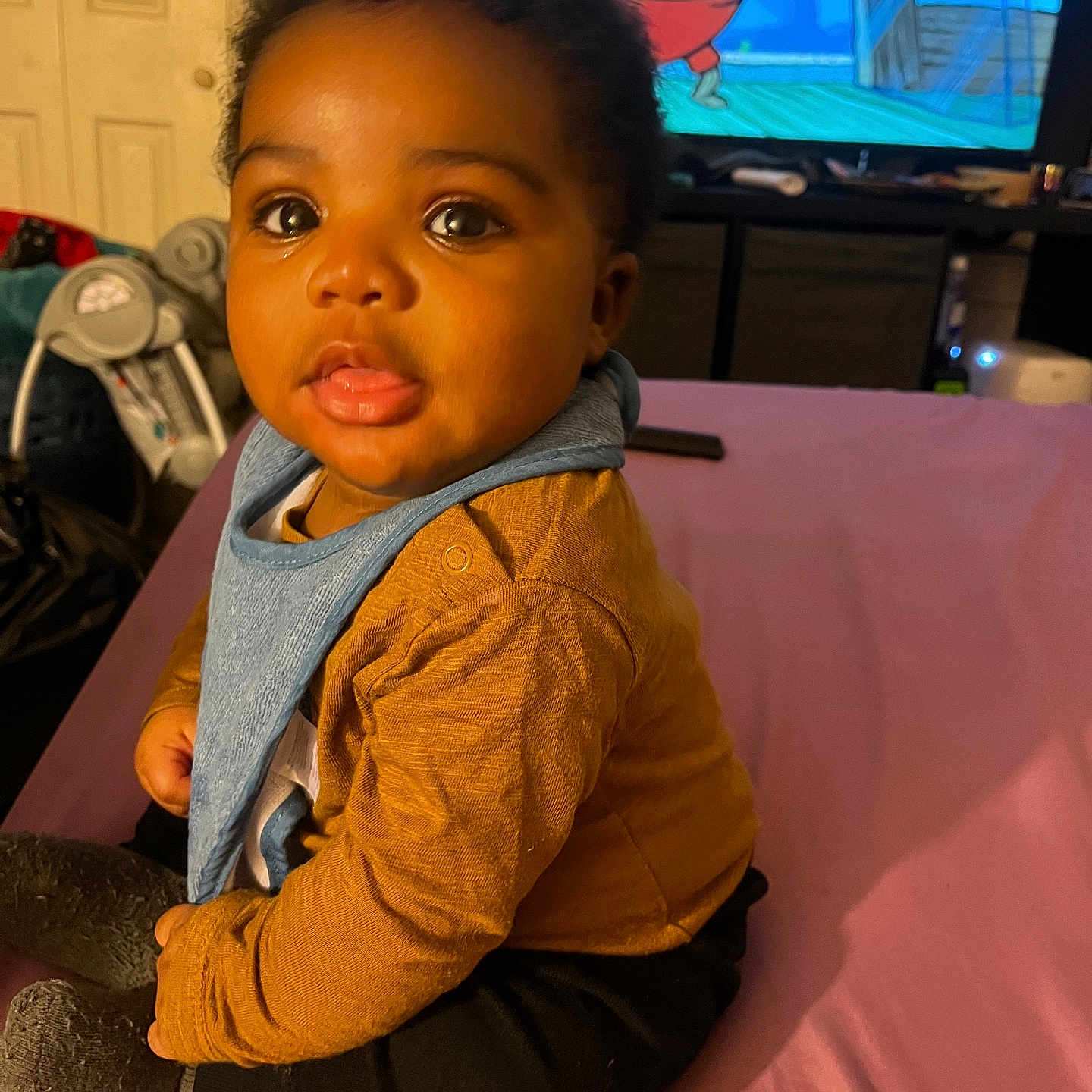 Messiah is registered to the contest to win money with this photo: baby, bib, black_pants, cartoon, child, clothing, curious, cute, eyes, face, furniture, hair, indoor, infant, mustard_shirt, pink_surface, portrait, room, sitting, television