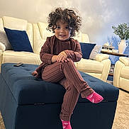 Valentina joined the competition — help win amazing prizes! toddler, child, curly_hair, smiling, socks, brown_outfit, ottoman, living_room, sofa, cushions, carpet, table, vase, books, remote_control, person, indoor, furniture, home_decor, happy