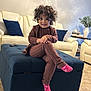 toddler, child, curly_hair, smiling, socks, brown_outfit, ottoman, living_room, sofa, cushions, carpet, table, vase, books, remote_control, person, indoor, furniture, home_decor, happy
