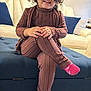 Valentina is registered to the contest to win money with this photo: toddler, child, smiling, curly_hair, brown_outfit, pink_socks, sitting, blue_ottoman, living_room, couch, pillows, carpet, happy, portrait, indoor, furniture, cozy, cute, person, crossed_legs