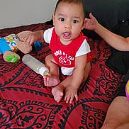 Jaycob joined the competition — help win amazing prizes! baby, child, infant, bed, bottle, toy, stuffed_animal, red_shirt, patterned_bedspread, hand, sitting, indoors, person, face, foot, arm, blanket, expression, casual, home