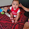 baby, child, infant, bed, bottle, toy, stuffed_animal, red_shirt, patterned_bedspread, hand, sitting, indoors, person, face, foot, arm, blanket, expression, casual, home