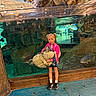 child, girl, stuffed_turtle, aquarium, fish, glass, indoor, reflection, rocks, water, floor, blue_floor, pigtails, pink_jacket, shoes, white_socks, backpack, serious_expression, large_toy, museum