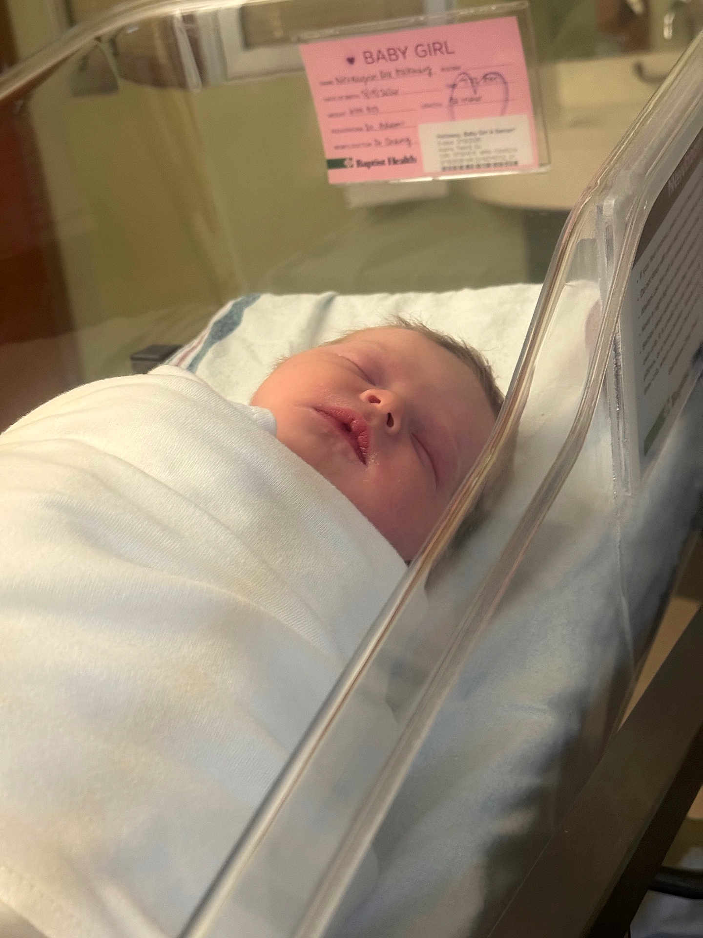 Novalynn is registered to the contest to win money with this photo: baby, newborn, infant, swaddled, sleeping, bassinet, hospital, crib, blanket, plastic, id_card, pink_sign, face, head, lips, peaceful, closeup, portrait, medical, nursery