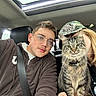 cat, tabby_cat, person, glasses, hoodie, cap, car_interior, seatbelt, sunroof, fluffy_cat, pet, animal, portrait, casual_clothing, friendship, selfie, indoor, feline, happy, companionship
