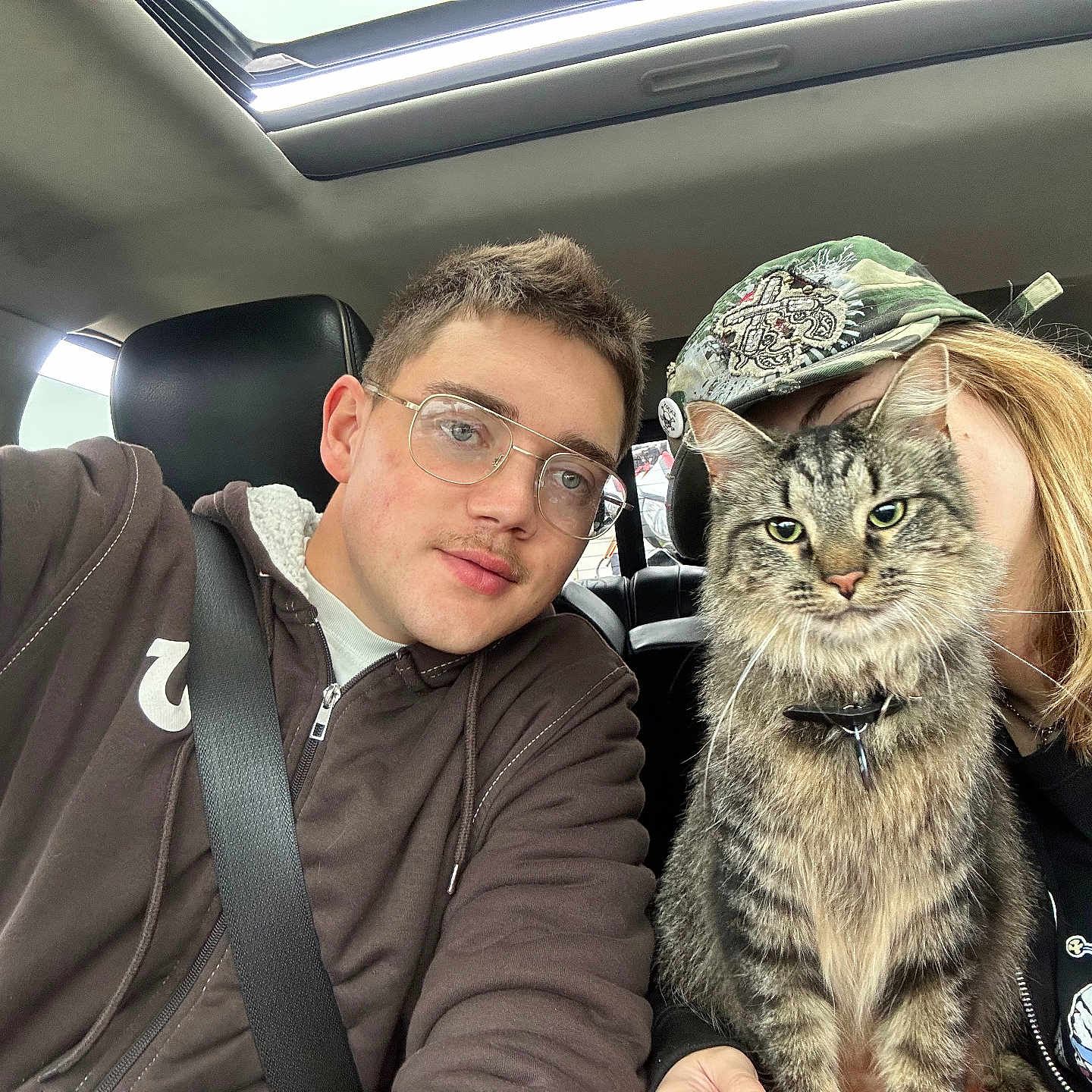 Stevie is registered to the contest to win money with this photo: cat, tabby_cat, person, glasses, hoodie, cap, car_interior, seatbelt, sunroof, fluffy_cat, pet, animal, portrait, casual_clothing, friendship, selfie, indoor, feline, happy, companionship