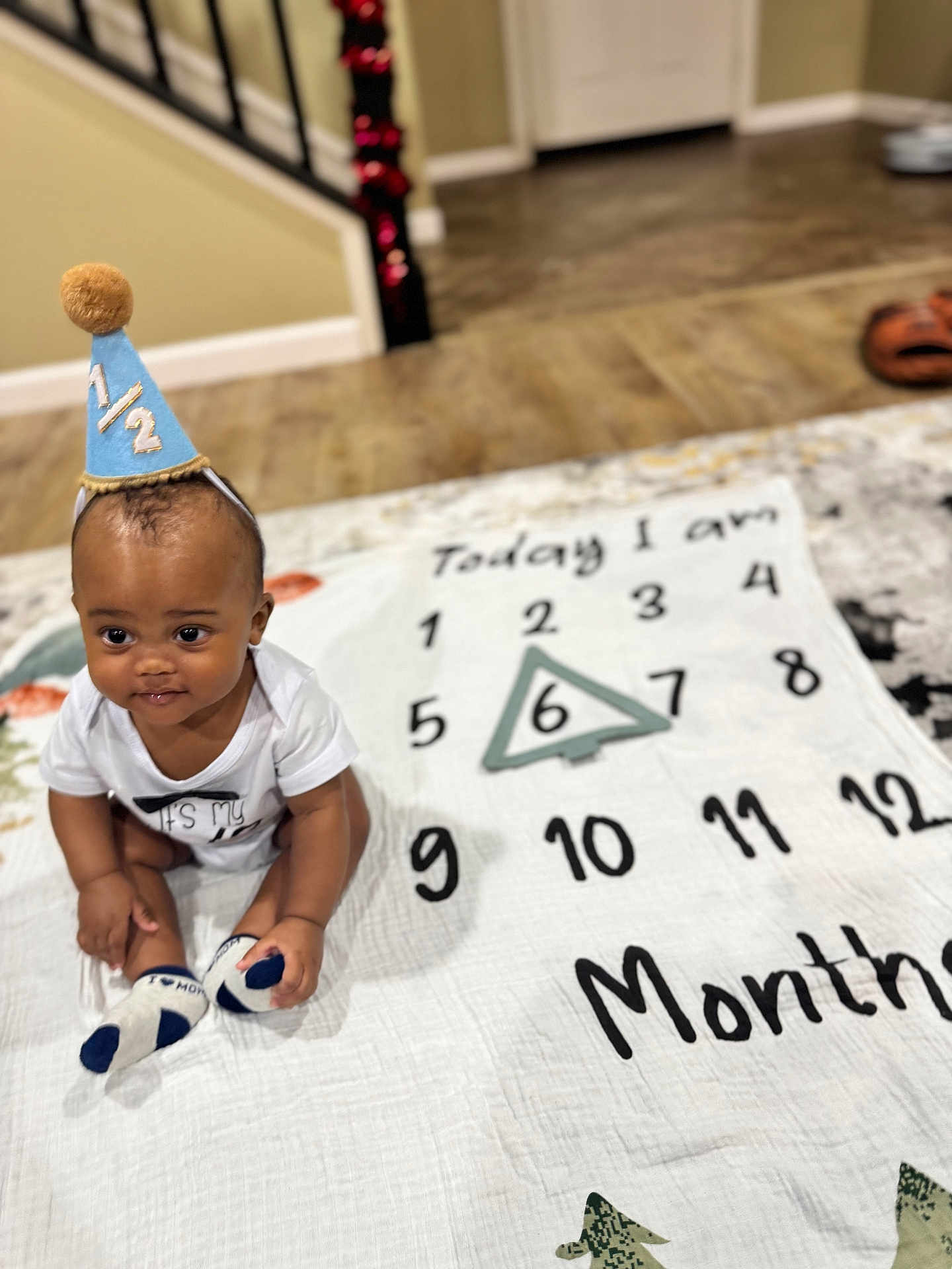 Kylen is registered to the contest to win money with this photo: baby, infant, six_months, milestone_blanket, party_hat, sitting, onesie, socks, indoor, hardwood_floor, portrait, smile, numbers, triangle_marker, celebration, staircase, decorations, photography, playtime, cute