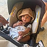 baby, child, car_seat, hat, blanket, infant, smiling, person, clothing, footwear, hand, outdoor, concrete_floor, casual, cute, seatbelt, headwear, toddler, young_child, sunlight