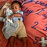 baby, child, infant, toddler, blanket, stuffed_animal, toy, clothing, shorts, shirt, curly_hair, lying_down, carpet, floor, indoor, person, cute, soft_toy, playtime, relaxing