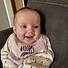 Murphy is registered to the contest to win money with this photo: baby, smiling, infant, bib, clothing, floral_pattern, couch, indoor, happy, cute, person, child, face, head, sitting, pink, fabric, skin, eyes, young