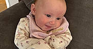Murphy joined the competition — help win amazing prizes! baby, infant, smiling, pink_pants, floral_clothing, bib, couch, brown_furniture, indoor, child, cute, happy, person, seated, head, face, small_child, cozy, home, casual