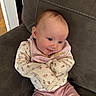 baby, infant, smiling, pink_pants, floral_clothing, bib, couch, brown_furniture, indoor, child, cute, happy, person, seated, head, face, small_child, cozy, home, casual