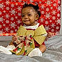 baby, child, smile, curly_hair, socks, red_outfit, gold_bow, christmas_ribbon, holiday, snowflake_pattern, red_background, gray_blanket, festive, indoors, portrait, cute, sitting, happy, celebration, person