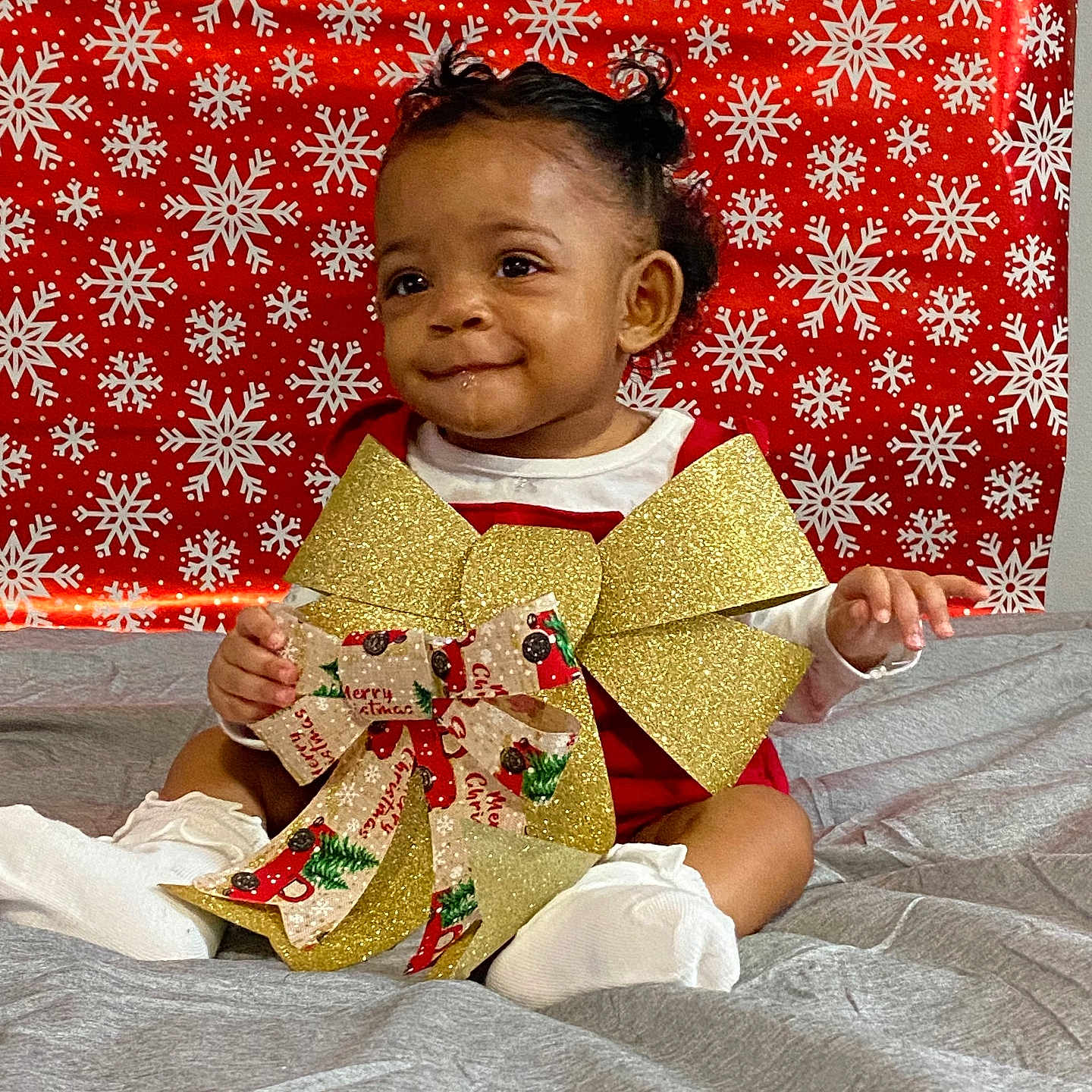 Z'Yane joined the competition — help win amazing prizes! baby, celebration, child, christmas_ribbon, curly_hair, cute, festive, gold_bow, gray_blanket, happy, holiday, indoors, person, portrait, red_background, red_outfit, sitting, smile, snowflake_pattern, socks