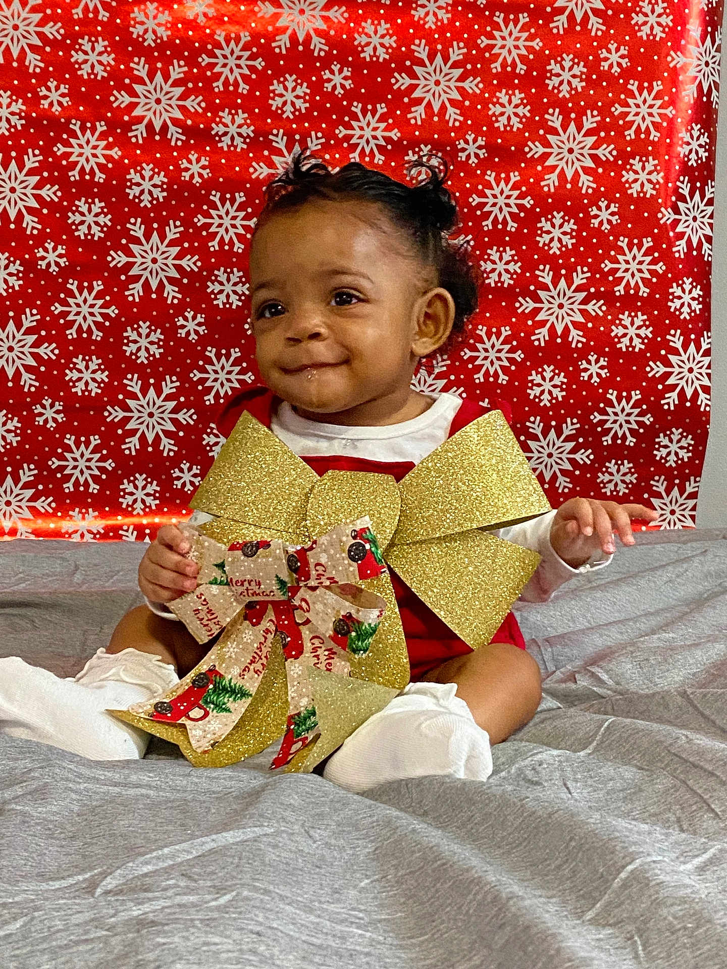 Z'Yane joined the competition — help win amazing prizes! baby, child, smile, curly_hair, socks, red_outfit, gold_bow, christmas_ribbon, holiday, snowflake_pattern, red_background, gray_blanket, festive, indoors, portrait, cute, sitting, happy, celebration, person