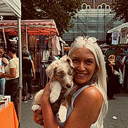 Maïko participe au concours pour gagner de l'argent avec cette photo : woman, puppy, dog, outdoor, market, stall, clothing, smile, white_hair, glasses, earrings, people, trees, daylight, street, casual, pet, happy, animal, shopping