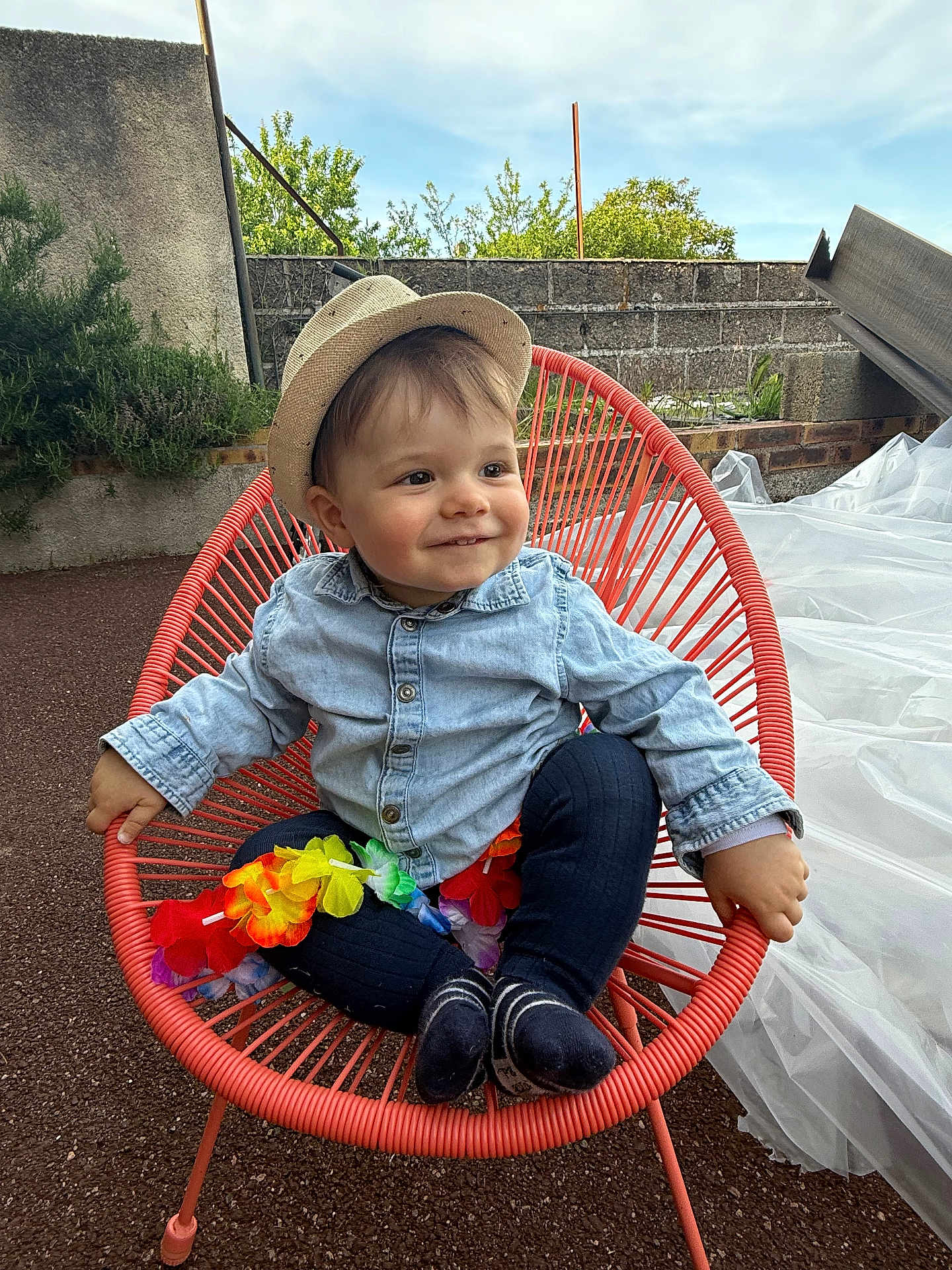 Léon a rejoint le concours — aidez-le/la à gagner de superbes lots ! toddler, child, hat, denim_shirt, orange_chair, flower_lei, outdoor, smiling, happy, cute, sitting, nature, daylight, face, person, grass, plants, blue_sky, playful, casual