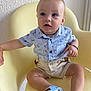 baby, child, blue_eyes, yellow_chair, light_blue_shirt, sailboat_print, beige_shorts, suspenders, blue_sandals, bear_prints, hand_holding, curious_expression, indoor, soft_lighting, portrait, cute, seated, person, footwear, casual_clothing