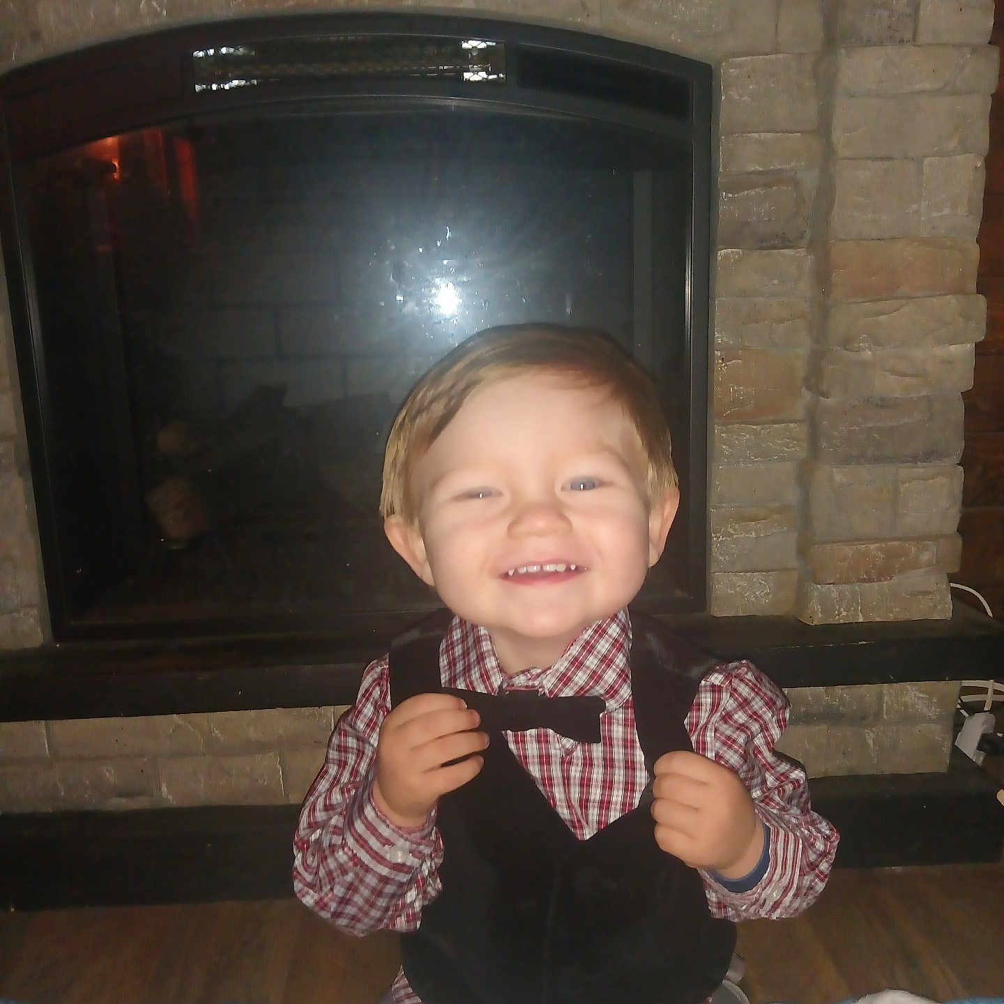 Alex is registered to the contest to win money with this photo: child, toddler, smile, happy, vest, bowtie, checkered_shirt, indoor, fireplace, stone_wall, blanket, floor, toy, baby, cute, portrait, clothing, sitting, flash, home